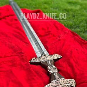 May include: A silver sword with a wooden handle and intricate carvings. The sword is lying on a red cloth with the text "BLAYDZ_KNIFE_CO" visible.