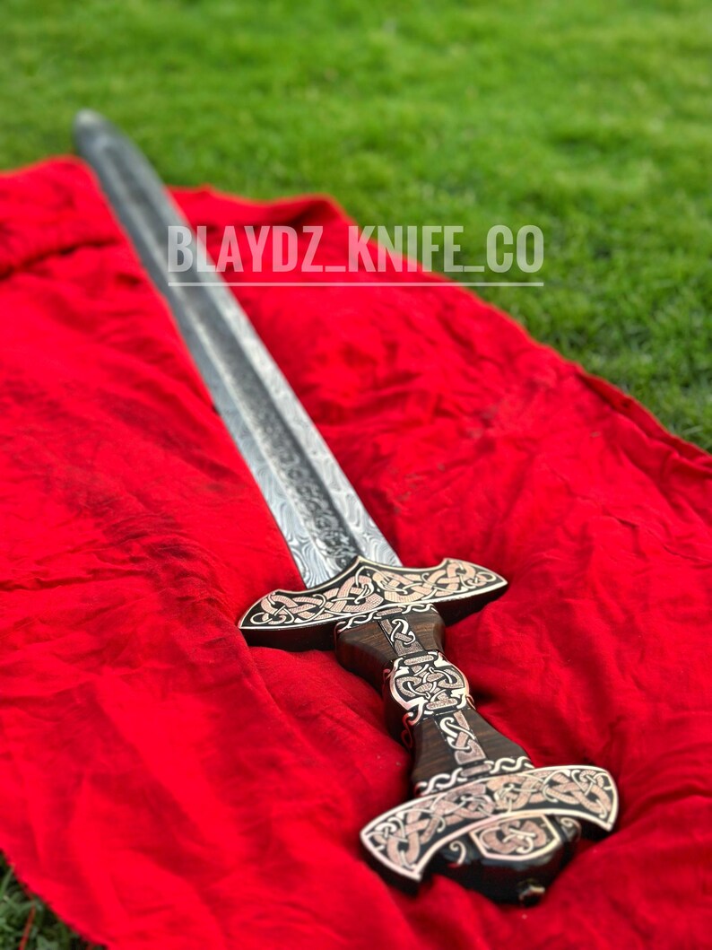May include: A silver sword with a wooden handle and intricate carvings. The sword is lying on a red cloth with the text "BLAYDZ_KNIFE_CO" visible.