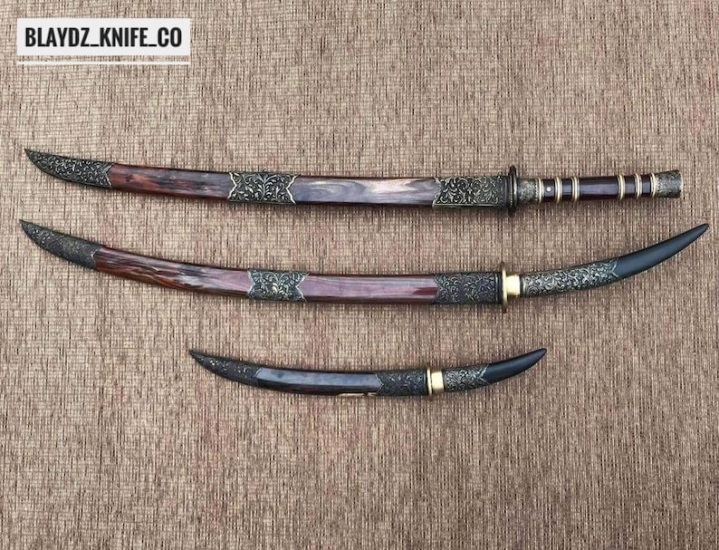 May include: Three decorative swords with ornate handles and blades. The swords have dark brown and black blades with decorative metal accents. The handles are a mix of dark brown and silver. The text "BLAYDZ_KNIFE_CO" is in the top left corner.