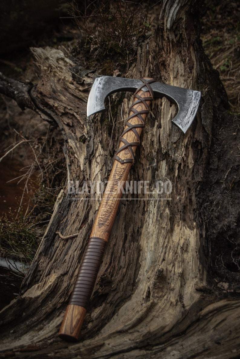 May include: A double-headed axe with a brown leather wrapped handle. The axe is resting on a tree stump with a rough, textured surface. The axe head is made of metal and has a hammered finish.