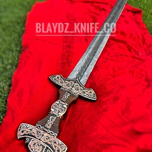 May include: A silver sword with a wooden handle and intricate metalwork. The sword has a Celtic knot design and is displayed on a red cloth. The text "BLAYDZ_KNIFE_CO" is visible on the cloth.
