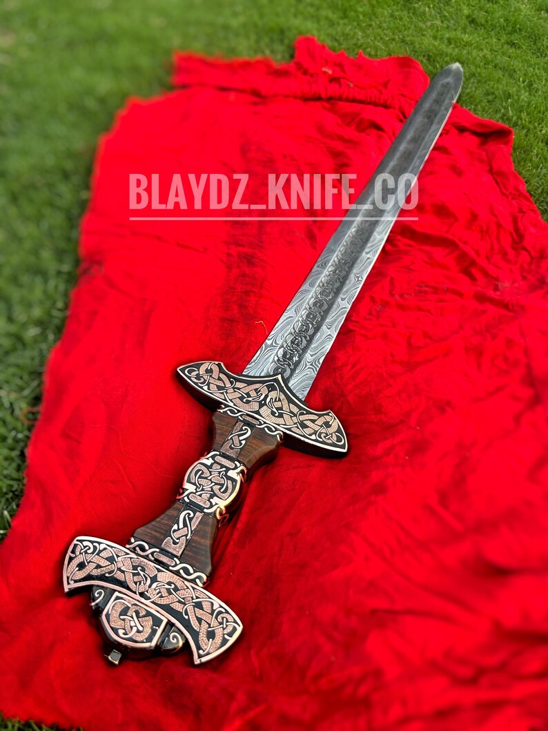 May include: A silver sword with a wooden handle and intricate metalwork. The sword has a Celtic knot design and is displayed on a red cloth. The text "BLAYDZ_KNIFE_CO" is visible on the cloth.
