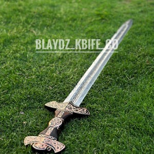May include: A silver sword with a black and gold handle, lying on a green grassy surface. The handle is decorated with intricate designs. The text "BLAYDZ_KBIFE_CO" is visible on the blade.