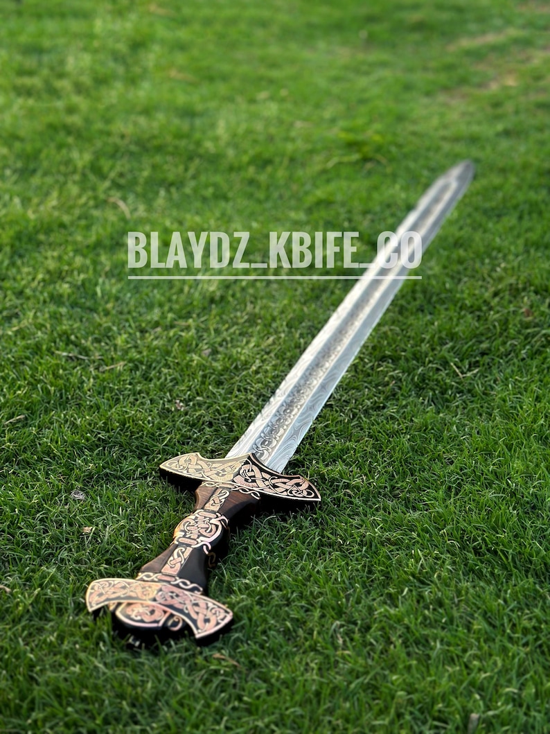 May include: A silver sword with a black and gold handle, lying on a green grassy surface. The handle is decorated with intricate designs. The text "BLAYDZ_KBIFE_CO" is visible on the blade.