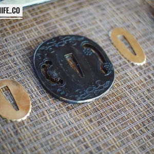 May include: A close-up of Japanese sword fittings, including a black tsuba with intricate designs and two smaller gold-coloured seppa. The tsuba features a central opening and decorative patterns. The text "BLAYDZ_KNIFE_CO" is in the top left corner.