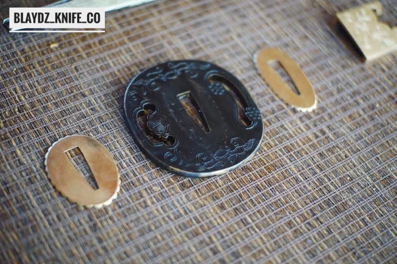 May include: A close-up of Japanese sword fittings, including a black tsuba with intricate designs and two smaller gold-coloured seppa. The tsuba features a central opening and decorative patterns. The text "BLAYDZ_KNIFE_CO" is in the top left corner.