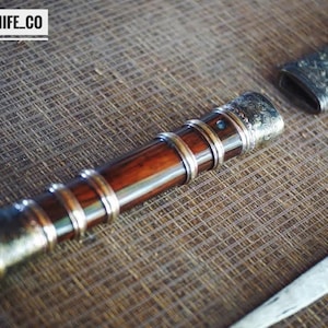 May include: A handcrafted knife with a wooden handle and metal detailing. The handle is a deep brown, with silver-coloured bands and end caps. The blade is partially visible, and the text "BLAYDZ_KNIFE_CO" is in the top left corner.