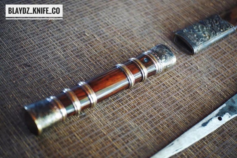 May include: A handcrafted knife with a wooden handle and metal detailing. The handle is a deep brown, with silver-coloured bands and end caps. The blade is partially visible, and the text "BLAYDZ_KNIFE_CO" is in the top left corner.