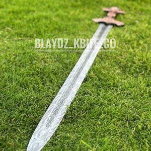 May include: A silver sword with a patterned blade and a brown wooden handle. The sword is lying on a bed of green grass.