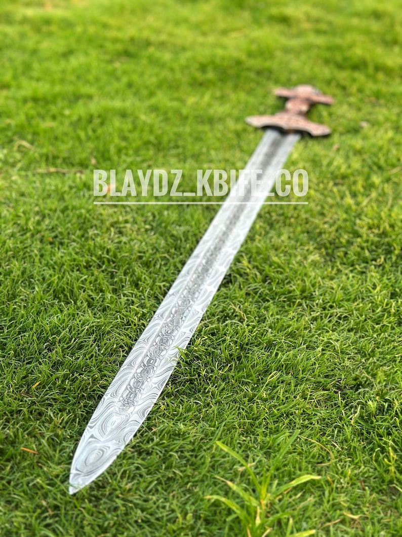 May include: A silver sword with a patterned blade and a brown wooden handle. The sword is lying on a bed of green grass.