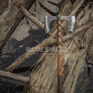 May include: A double-headed axe with a brown leather handle and a wooden shaft. The axe is resting on a log with a body of water in the background. The axe has a polished finish and the handle is wrapped in leather with a braided design.