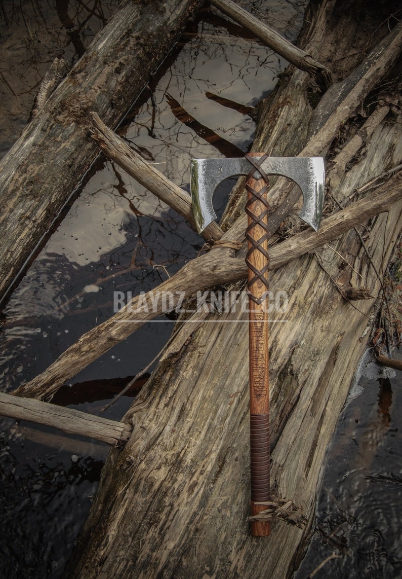 May include: A double-headed axe with a brown leather handle and a wooden shaft. The axe is resting on a log with a body of water in the background. The axe has a polished finish and the handle is wrapped in leather with a braided design.