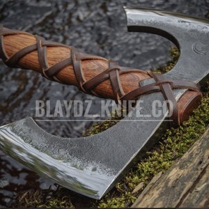 May include: A double-bit axe with a wooden handle wrapped in brown leather. The axe head is made of metal and has a polished finish. The axe is resting on a piece of wood covered in moss. The text "BLAYDZ KNIFE CO" is visible on the wood.