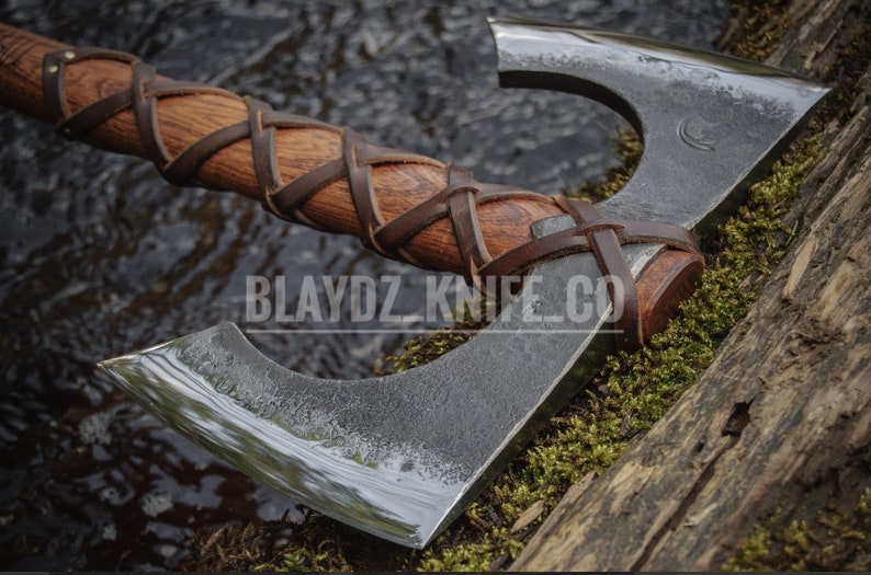 May include: A double-bit axe with a wooden handle wrapped in brown leather. The axe head is made of metal and has a polished finish. The axe is resting on a piece of wood covered in moss. The text "BLAYDZ KNIFE CO" is visible on the wood.