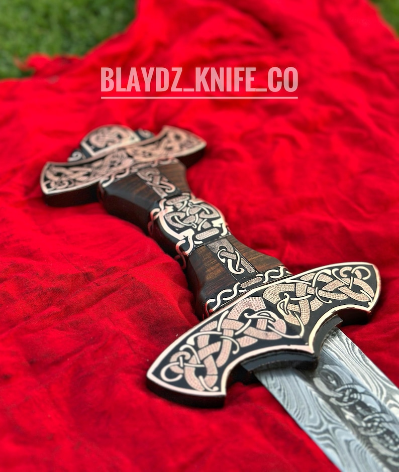 May include: A decorative sword with a wooden handle and a silver blade. The handle is intricately carved with a Celtic knot design and the blade is Damascus steel. The sword is lying on a red cloth with the text "BLAYDZ_KNIFE_CO" visible in the background.