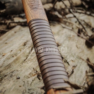 May include: Close-up of a wooden axe handle with a brown leather wrap and intricate carvings. The handle is resting on a piece of wood with the text "BLAYDZ KNIFE CO." visible.