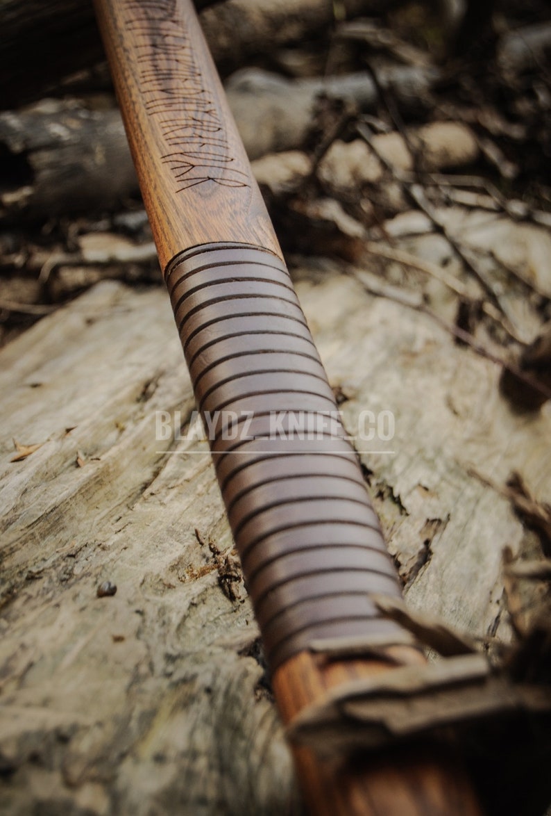 May include: Close-up of a wooden axe handle with a brown leather wrap and intricate carvings. The handle is resting on a piece of wood with the text "BLAYDZ KNIFE CO." visible.