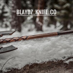 May include: A double-headed axe with a wooden handle wrapped in leather. The axe is lying on a bed of snow with a blurred background of trees. The text "BLAYDZ_KNIFE_CO" is visible in the image.