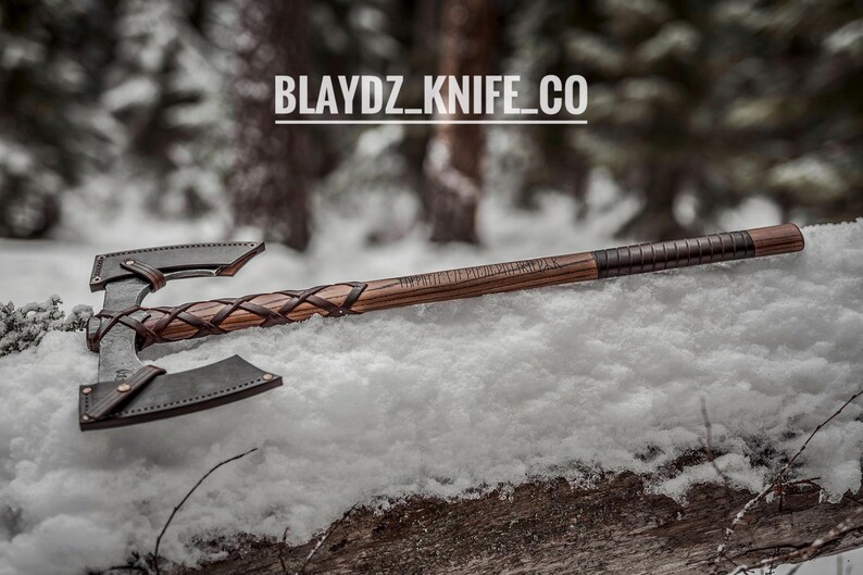 May include: A double-headed axe with a wooden handle wrapped in leather. The axe is lying on a bed of snow with a blurred background of trees. The text "BLAYDZ_KNIFE_CO" is visible in the image.