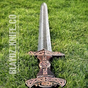 May include: A silver sword with a dark wood handle and intricate gold designs. The sword is lying on a bed of green grass.