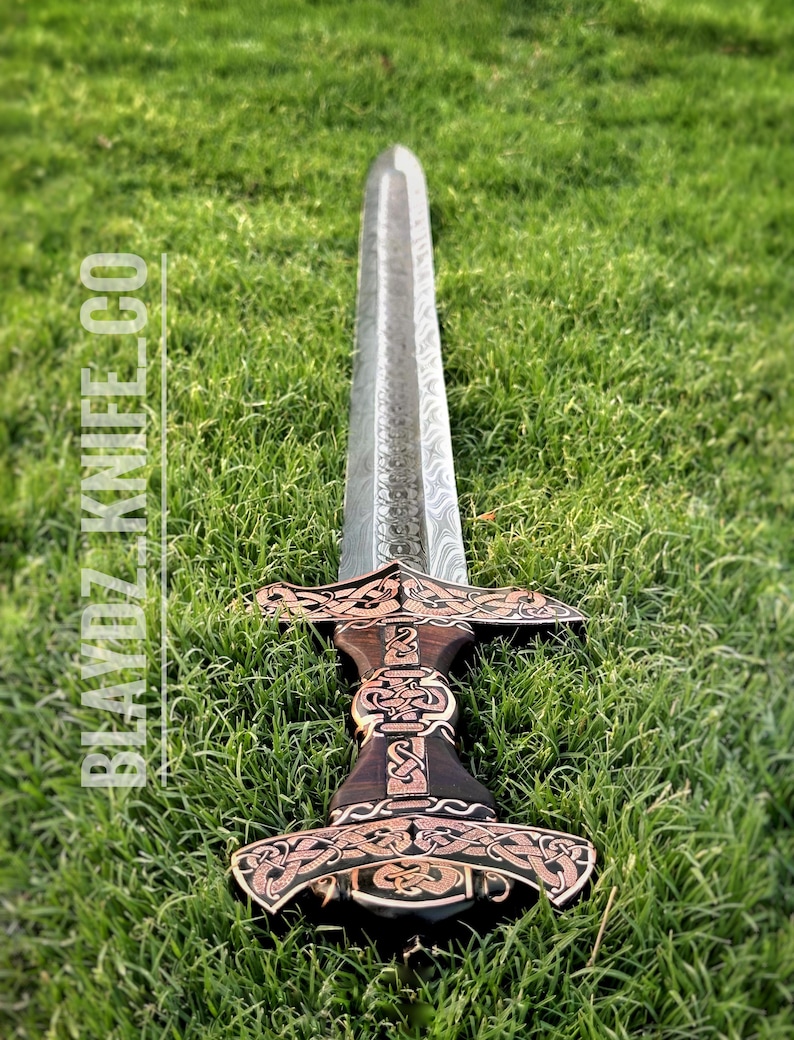 May include: A silver sword with a dark wood handle and intricate gold designs. The sword is lying on a bed of green grass.