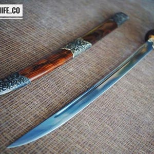 May include: A handcrafted sword with a polished steel blade and a wooden handle. The sword is accompanied by a wooden sheath with decorative metal accents. The text "BLAYDZ_KNIFE_CO" is visible in the upper left corner.