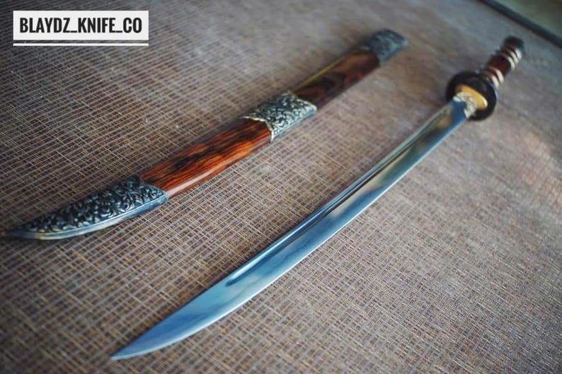 May include: A handcrafted sword with a polished steel blade and a wooden handle. The sword is accompanied by a wooden sheath with decorative metal accents. The text "BLAYDZ_KNIFE_CO" is visible in the upper left corner.