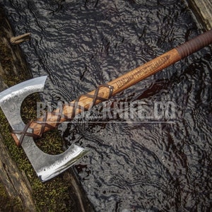 May include: A double-bit axe with a wooden handle wrapped in brown leather. The axe is lying on a wooden surface next to a stream.