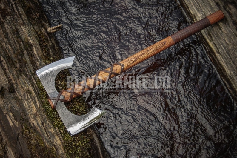 May include: A double-bit axe with a wooden handle wrapped in brown leather. The axe is lying on a wooden surface next to a stream.