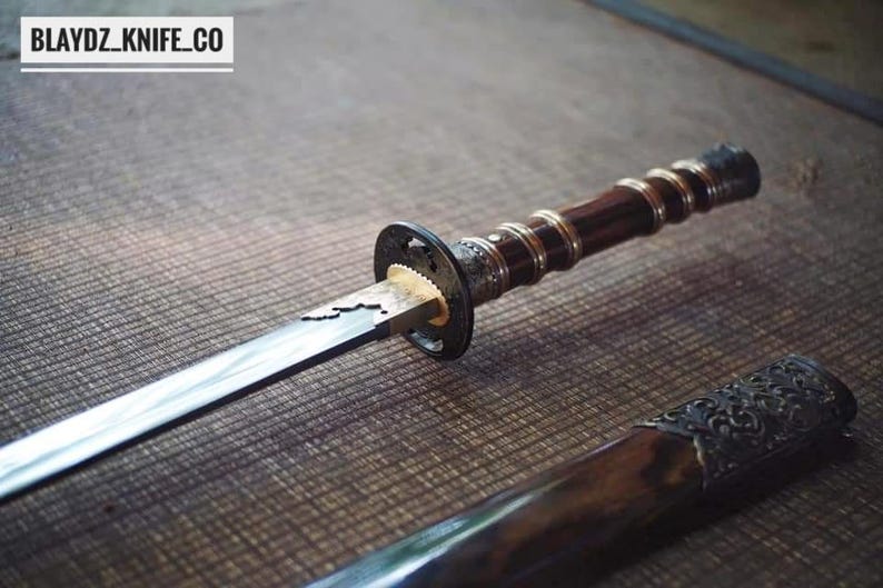 May include: A Japanese-style sword with a dark wooden handle and a silver blade. The handle has a detailed design with a dark brown finish. The scabbard is visible at the bottom. The text "BLAYDZ_KNIFE_CO" is at the top.