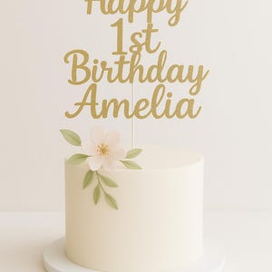 May include: A white iced cake with a gold glitter cake topper reading "Happy 1st Birthday Amelia". The cake is decorated with a pale pink flower and green leaves. The cake sits on a white cake stand.