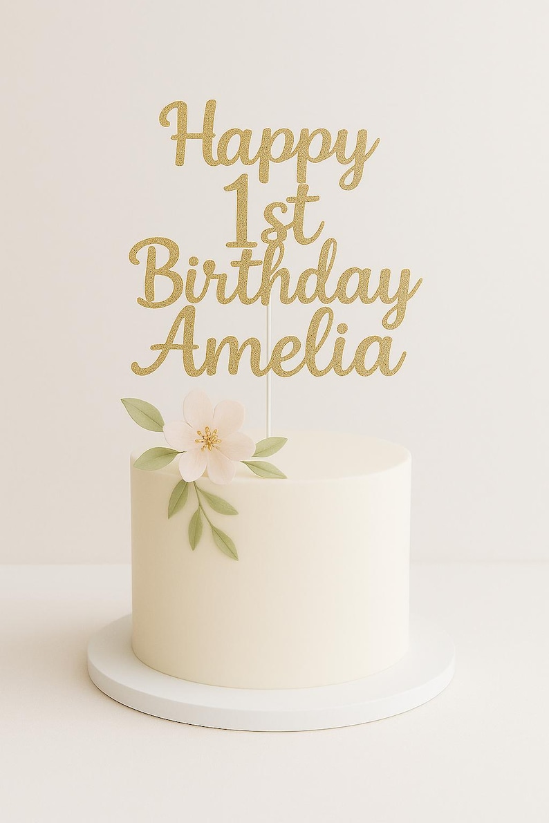 May include: A white iced cake with a gold glitter cake topper reading "Happy 1st Birthday Amelia". The cake is decorated with a pale pink flower and green leaves. The cake sits on a white cake stand.