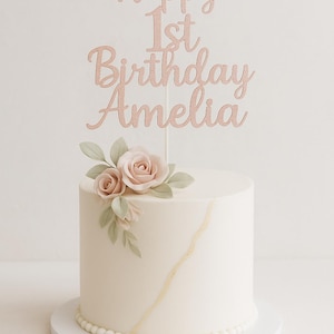 May include: A white cake with a gold design and a floral arrangement. The cake is topped with a rose gold cake topper that reads "Happy 1st Birthday Amelia". The cake sits on a white cake board.