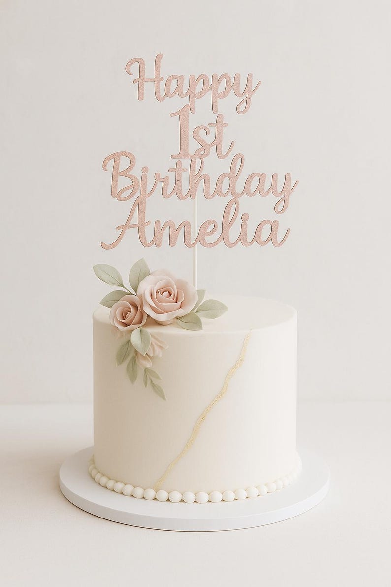 May include: A white cake with a gold design and a floral arrangement. The cake is topped with a rose gold cake topper that reads "Happy 1st Birthday Amelia". The cake sits on a white cake board.