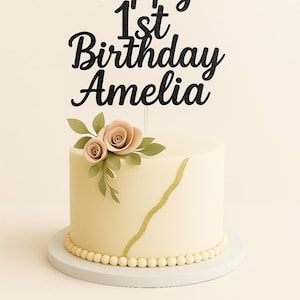 May include: A cream-coloured cake with a gold stripe and pearl detailing. A black glitter cake topper reads "Happy 1st Birthday Amelia". The cake is decorated with peach-coloured roses and green leaves. The cake sits on a white cake stand.