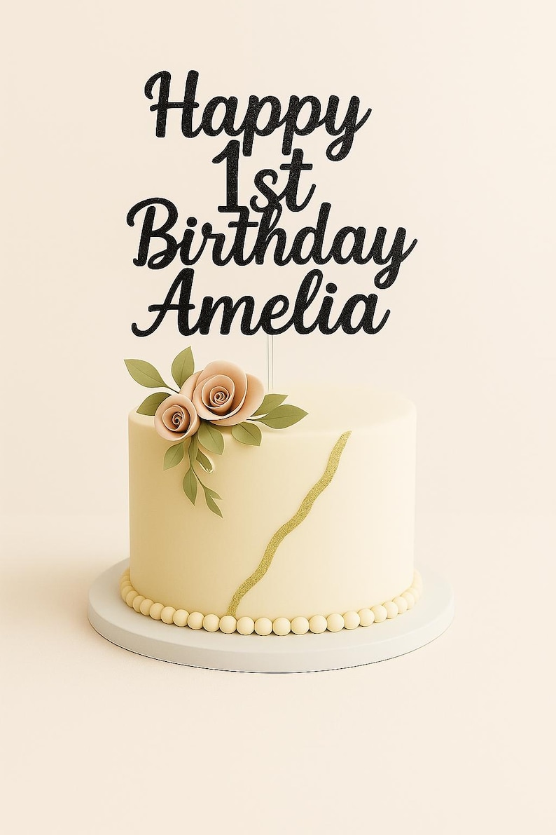 May include: A cream-coloured cake with a gold stripe and pearl detailing. A black glitter cake topper reads "Happy 1st Birthday Amelia". The cake is decorated with peach-coloured roses and green leaves. The cake sits on a white cake stand.
