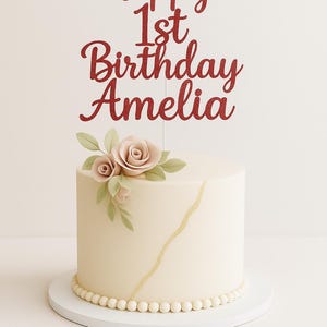 May include: A cream-coloured cake with a gold stripe and a beaded base. It's topped with a red glitter "Happy 1st Birthday Amelia" cake topper and floral decorations. The cake sits on a white cake stand. The cake is round.
