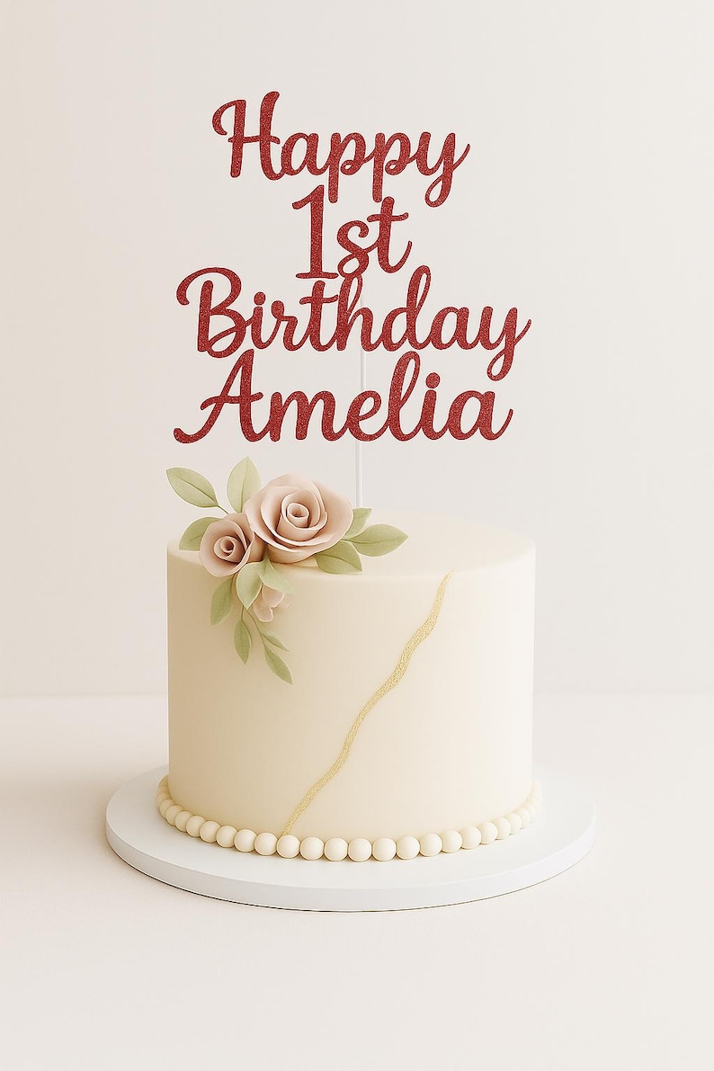 May include: A cream-coloured cake with a gold stripe and a beaded base. It's topped with a red glitter "Happy 1st Birthday Amelia" cake topper and floral decorations. The cake sits on a white cake stand. The cake is round.