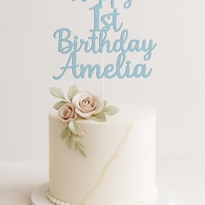 May include: A round, white frosted cake with a light blue cake topper that reads "Happy 1st Birthday Amelia." The cake is decorated with two pale pink roses and green leaves. A gold line accents the side, and the base is lined with white pearls.
