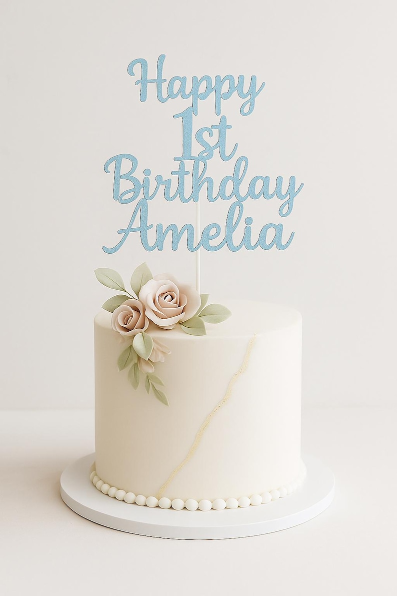 May include: A round, white frosted cake with a light blue cake topper that reads "Happy 1st Birthday Amelia." The cake is decorated with two pale pink roses and green leaves. A gold line accents the side, and the base is lined with white pearls.