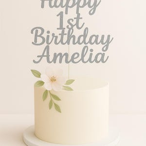 May include: A round, white frosted cake with a silver glitter cake topper that reads "Happy 1st Birthday Amelia." The cake is decorated with a white flower and green leaves. The cake sits on a white cake stand.