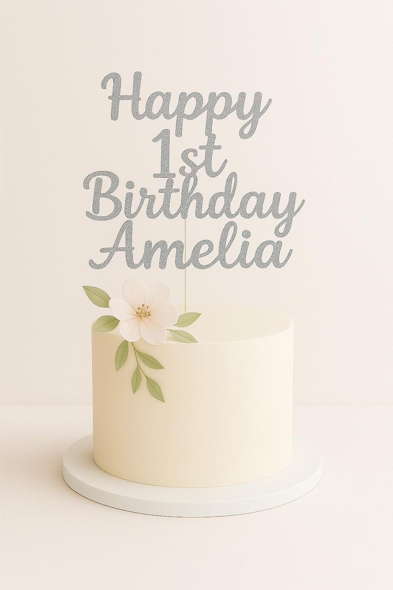 May include: A round, white frosted cake with a silver glitter cake topper that reads "Happy 1st Birthday Amelia." The cake is decorated with a white flower and green leaves. The cake sits on a white cake stand.