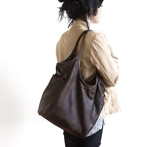 May include: A brown leather tote bag with a large pocket on the front. The bag is worn over the shoulder of a person wearing a beige jacket.