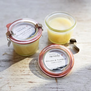 May include: Three small glass jars filled with a yellow balm. Two jars have red-rimmed lids with metal closures and labels that read "leather remedy + ROUGH & TUMBLE made in maine." The third jar is open.