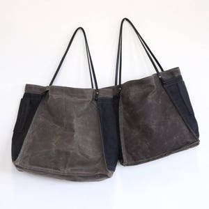 May include: Two large canvas tote bags with black leather handles. The bags are made of a dark brown canvas with a black canvas panel on the side. The bags are hanging on a white wall.