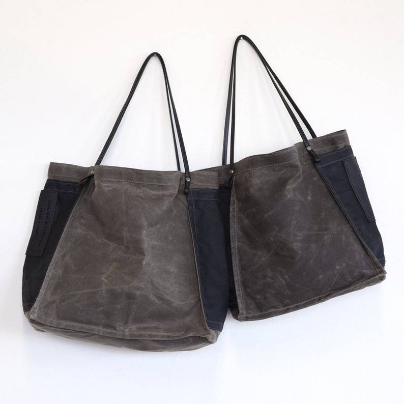 May include: Two large canvas tote bags with black leather handles. The bags are made of a dark brown canvas with a black canvas panel on the side. The bags are hanging on a white wall.