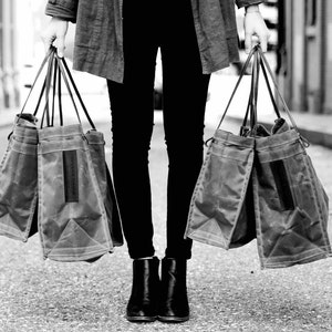 May include: A person carrying two large, gray canvas tote bags with leather handles. The bags are made of a durable, water-resistant material.