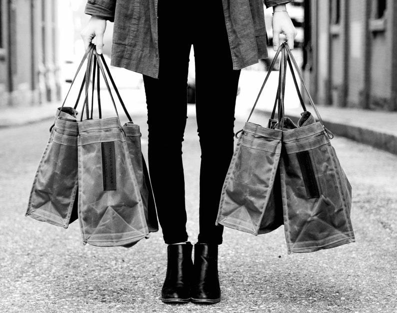 May include: A person carrying two large, gray canvas tote bags with leather handles. The bags are made of a durable, water-resistant material.