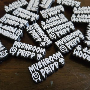 May include: A selection of black and white Mushroom Pripp charms. Each charm displays the text "Mushroom Pripp" in a distinctive font, alongside an Instagram logo and a mushroom graphic. The charms are arranged on a wooden surface.