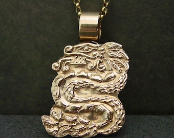 May include: A gold-toned metal pendant shaped like a coiled dragon with intricate scale and leaf detailing, suspended from a matching gold-toned chain. The pendant features a smooth, polished bail at the top, connecting the dragon figure to the chain.